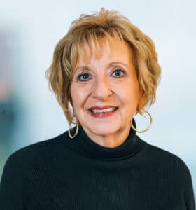 A woman with short blonde hair and hoop earrings smiles at the camera. She is wearing a black, long-sleeve top. The background is softly blurred.