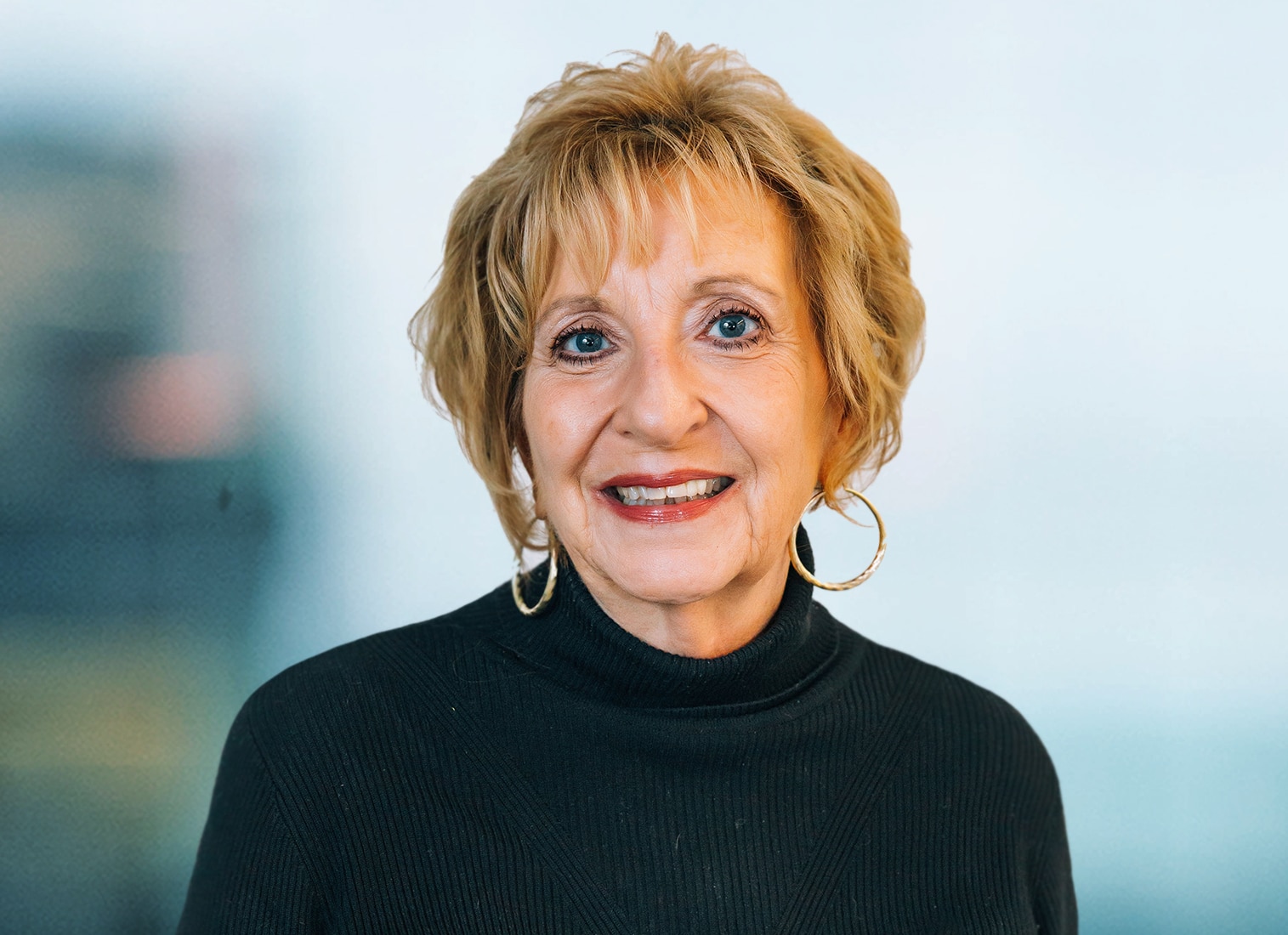 A woman with short blonde hair and hoop earrings smiles at the camera. She is wearing a black, long-sleeve top. The background is softly blurred.