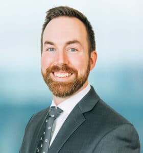 A man with a beard and mustache is smiling. He is wearing a dark suit, a white shirt, and a patterned tie. The background is a blurred light blue.