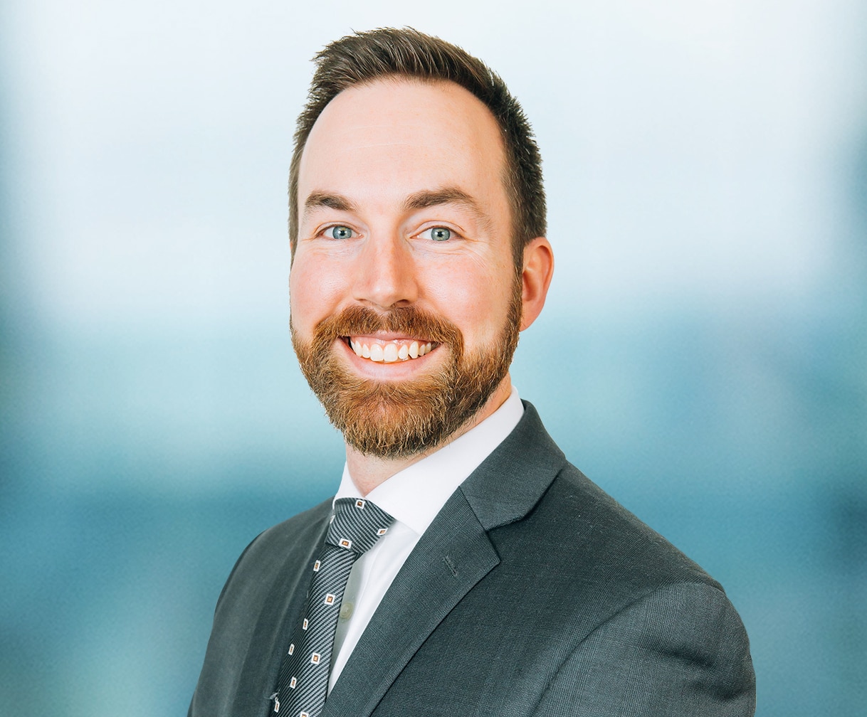 A man with a beard and mustache is smiling. He is wearing a dark suit, a white shirt, and a patterned tie. The background is a blurred light blue.