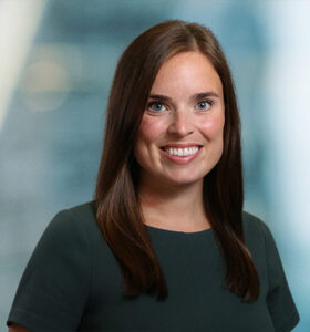 A woman with long brown hair wearing a dark green top smiles at the camera against a blurred blue and gray background.