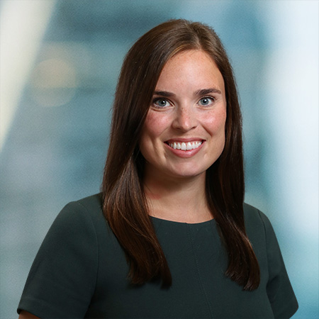 A woman with long brown hair wearing a dark green top smiles at the camera against a blurred blue and gray background.