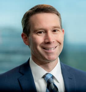 A man in a blue suit and checkered tie smiles at the camera. The background is blurry, showing a window with an abstract view of a cityscape.