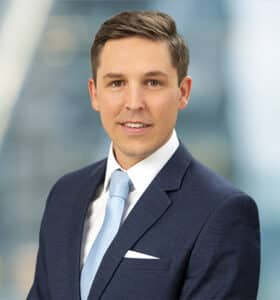 Professional man in a navy suit with a light blue tie stands against a blurred office background.