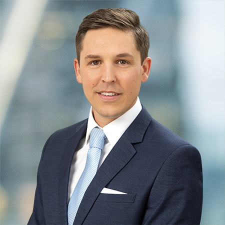 Professional man in a navy suit with a light blue tie stands against a blurred office background.
