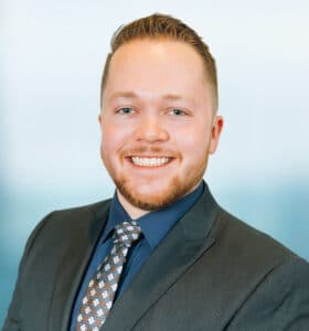A man with short hair and a beard is smiling, wearing a dark suit, navy shirt, and patterned tie. He is standing against a blurred blue and white background.