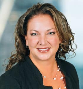 A woman with wavy brown hair smiles at the camera. She is wearing a black blazer and a pearl necklace. The background is blurred, suggesting an office or professional setting.
