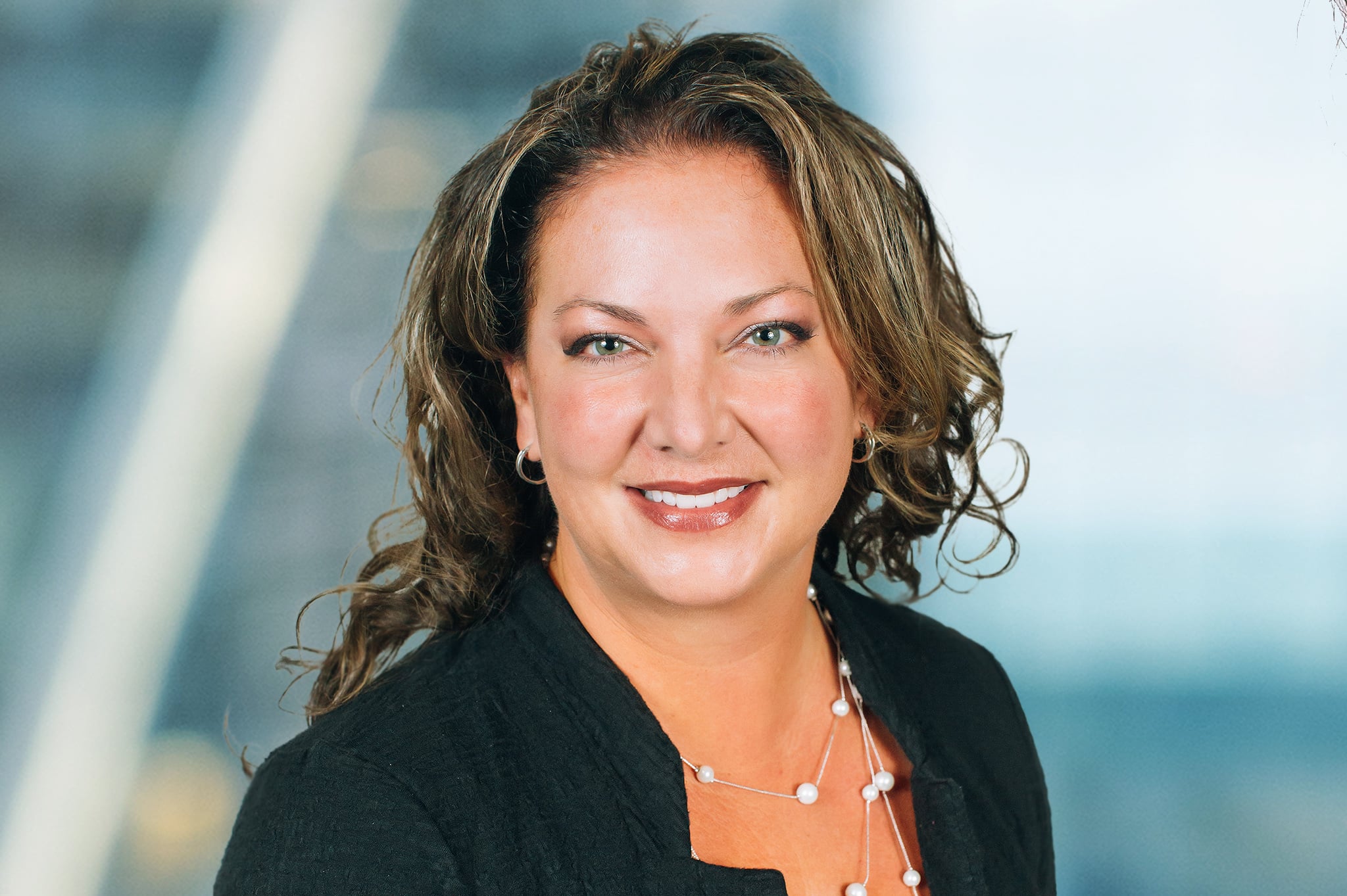 A woman with wavy brown hair smiles at the camera. She is wearing a black blazer and a pearl necklace. The background is blurred, suggesting an office or professional setting.