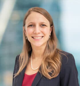 A person with long, light brown hair is smiling, wearing a dark blazer, a red top, and a pearl necklace. The background is blurred with a blue and white tone.