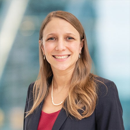A person with long, light brown hair is smiling, wearing a dark blazer, a red top, and a pearl necklace. The background is blurred with a blue and white tone.
