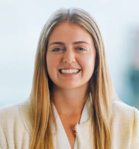 A person with long blonde hair smiles at the camera. They are wearing a white blazer, and the background is softly blurred, suggesting an indoor setting.