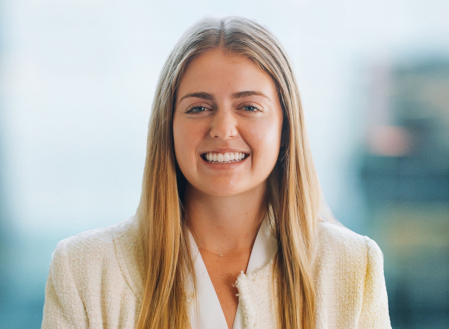 A person with long blonde hair smiles at the camera. They are wearing a white blazer, and the background is softly blurred, suggesting an indoor setting.