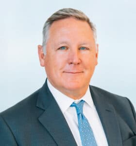 A middle-aged man wearing a suit and a light blue tie stands against a soft, blurred background. He has short, gray hair and a confident expression.