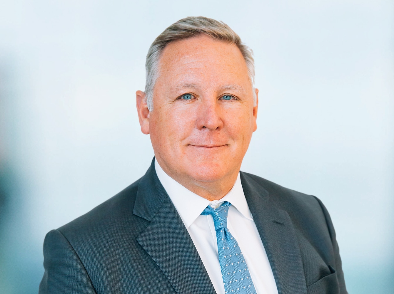 Sean-Cook-e1637184427301 A middle-aged man wearing a suit and a light blue tie stands against a soft, blurred background. He has short, gray hair and a confident expression.