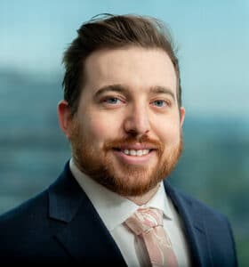 A man with a beard and styled hair is smiling at the camera. He is wearing a navy suit, a white shirt, and a light pink tie. The background is blurred, showing a cityscape through a window.