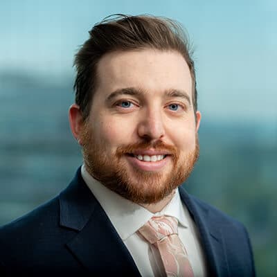 A man with a beard and styled hair is smiling at the camera. He is wearing a navy suit, a white shirt, and a light pink tie. The background is blurred, showing a cityscape through a window.