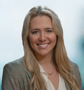 Smiling person with long blonde hair wearing a plaid blazer and a white blouse. The background is softly blurred.