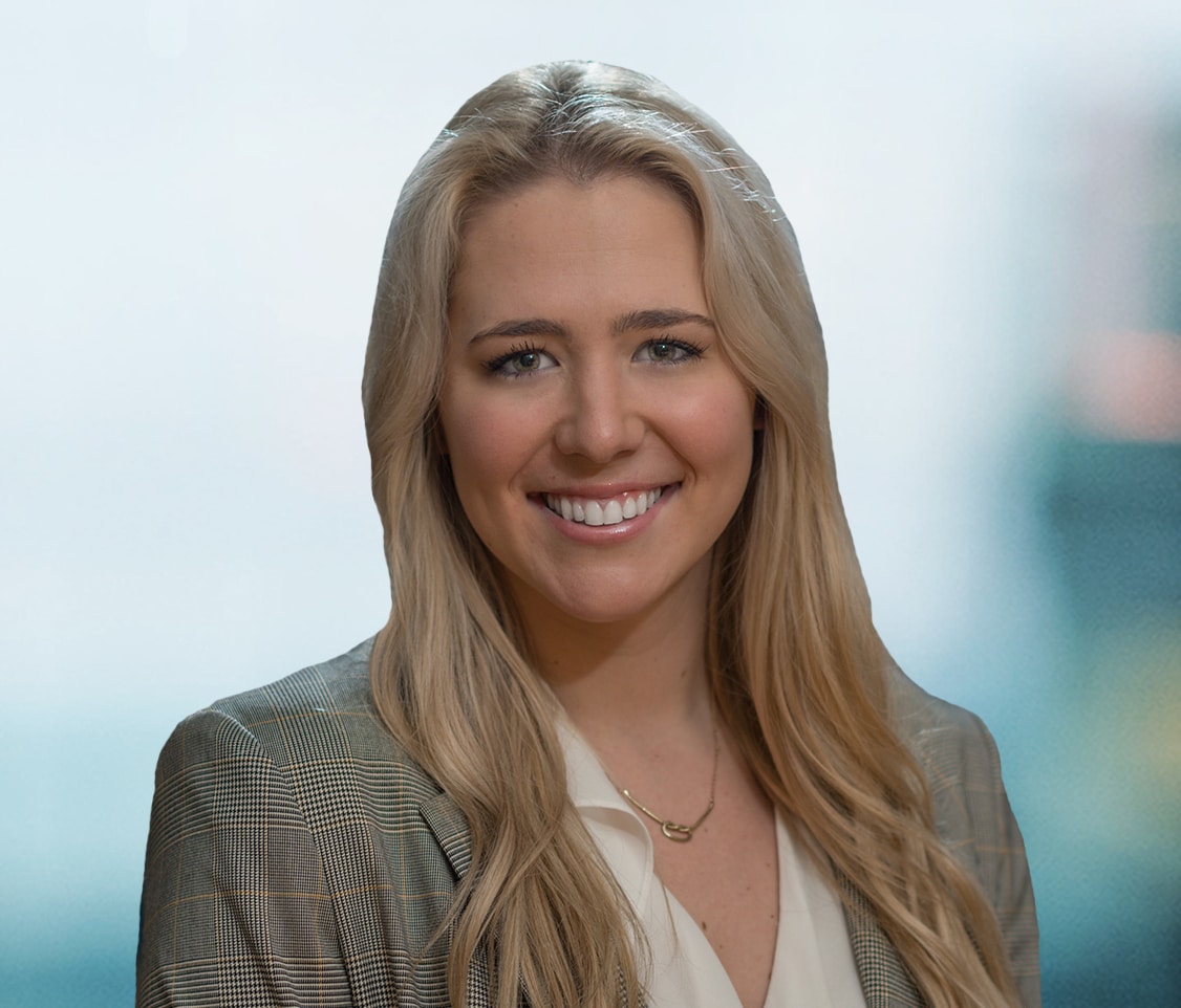 Smiling person with long blonde hair wearing a plaid blazer and a white blouse. The background is softly blurred.