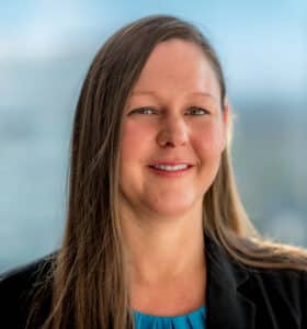 A woman with long brown hair smiles at the camera. She is wearing a black blazer over a blue top. The background is blurred, showing a window with a view of a cityscape on a bright day.