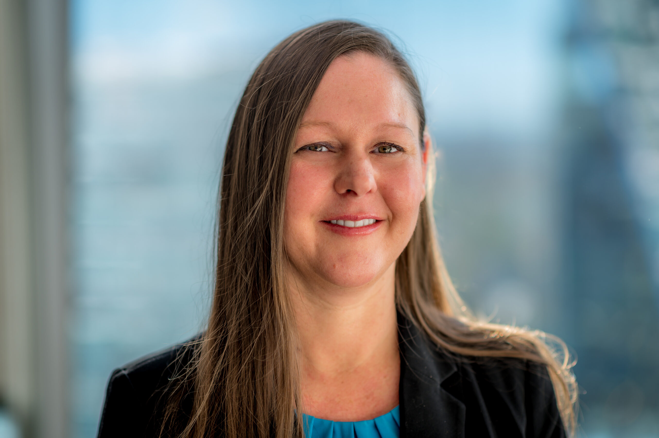Sheila-Bozeman A woman with long brown hair smiles at the camera. She is wearing a black blazer over a blue top. The background is blurred, showing a window with a view of a cityscape on a bright day.