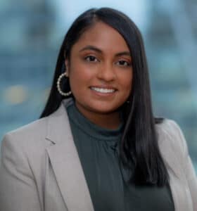 A person with long black hair wearing a beige blazer and green top smiles at the camera. Blurred office windows are visible in the background.
