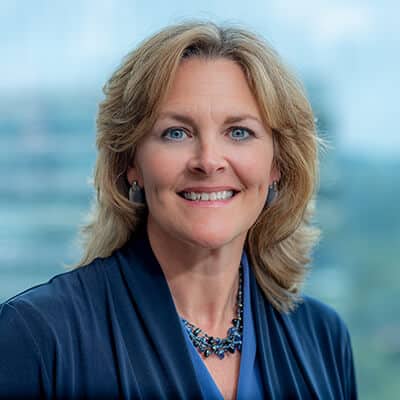 A woman with shoulder-length blonde hair smiles warmly. She is wearing a dark blue blouse and a matching beaded necklace. The background shows an out-of-focus cityscape through a large window.