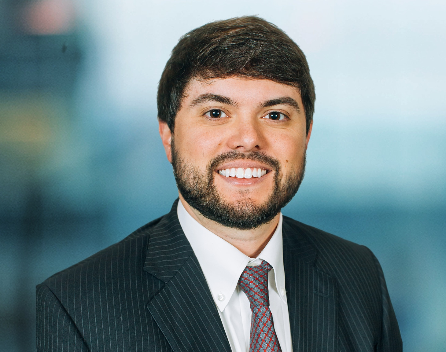 A man with short brown hair and a beard is smiling. He is wearing a dark pinstripe suit, white shirt, and a maroon patterned tie. The background is blurred, featuring shades of blue and gray.