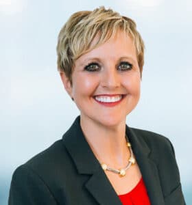 A person with short blonde hair is smiling, dressed in a black blazer over a red shirt, accessorized with a gold necklace. The background is softly blurred, featuring shades of blue and white.