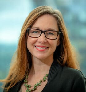 A woman with long brown hair and glasses smiles at the camera. She is wearing a black top and a green beaded necklace. The background is blurred, suggesting an outdoor or glass-enclosed setting.