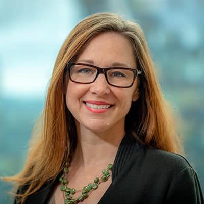 A woman with long brown hair and glasses smiles at the camera. She is wearing a black top and a green beaded necklace. The background is blurred, suggesting an outdoor or glass-enclosed setting.
