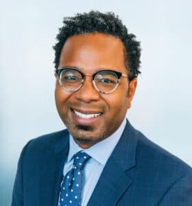 A person with curly hair and glasses is smiling at the camera, wearing a blue suit and a polka-dot tie. The background is a soft, blurred light blue.