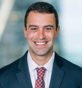 A man in a dark suit and red patterned tie smiles at the camera. The background is a blurred indoor setting with light streaming in.
