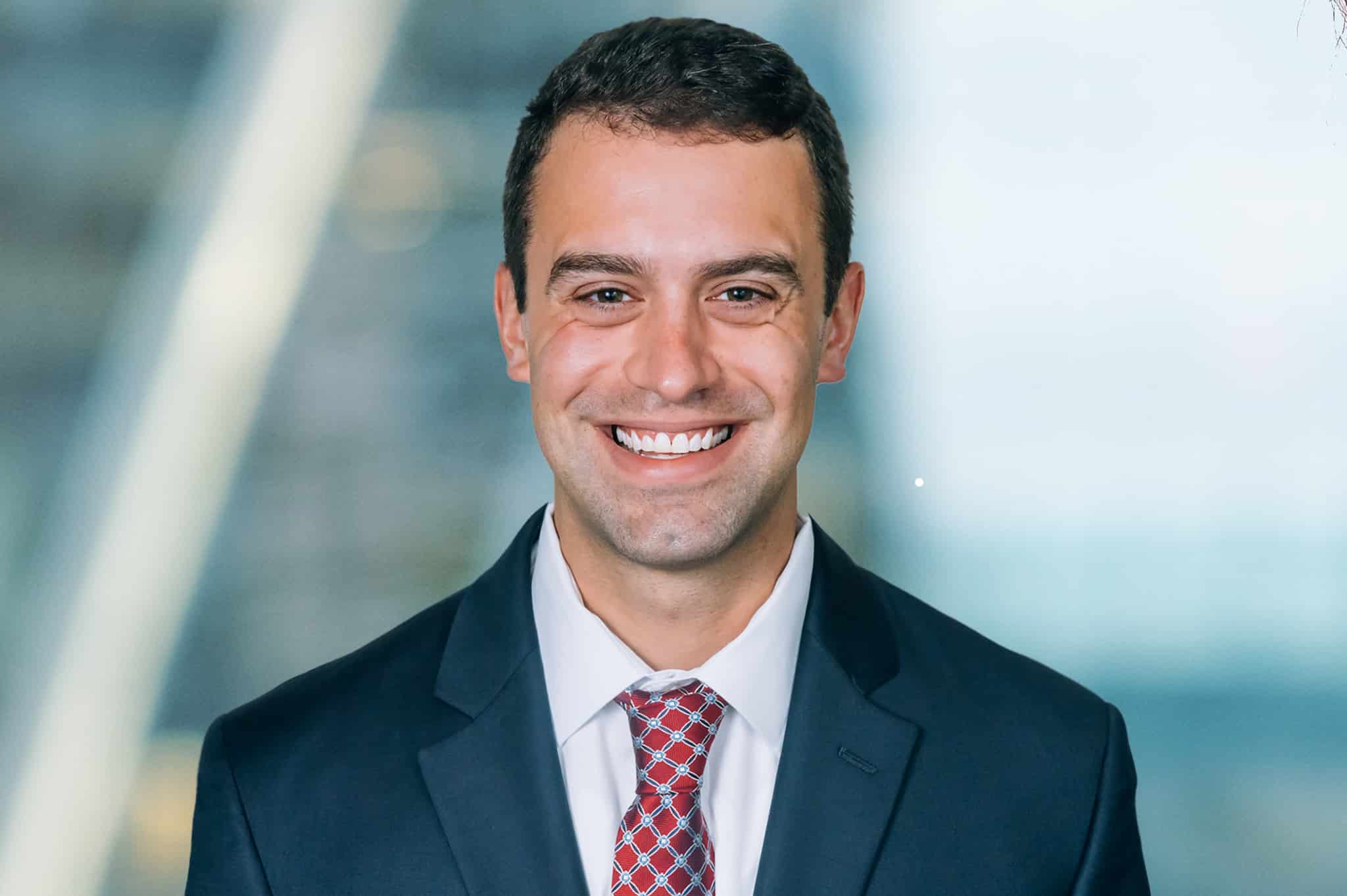 A man in a dark suit and red patterned tie smiles at the camera. The background is a blurred indoor setting with light streaming in.