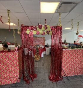 An office cubicle is festively decorated with red tinsel curtains, garlands, and colorful ornaments. WLY WOWDERS is spelled out in letters above the entrance. Holiday decorations include a snowman and Santa-themed wrapping paper.