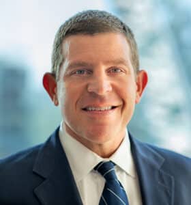 A man in a suit and tie smiles at the camera with a blurred cityscape in the background.