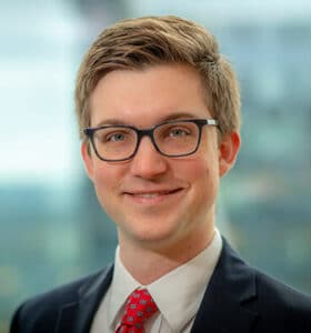 A person with short light brown hair, wearing glasses, a white shirt, a red tie with patterns, and a dark suit jacket, smiling at the camera. The background is blurred, suggesting an office or cityscape setting.