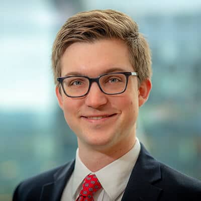 A person with short light brown hair, wearing glasses, a white shirt, a red tie with patterns, and a dark suit jacket, smiling at the camera. The background is blurred, suggesting an office or cityscape setting.