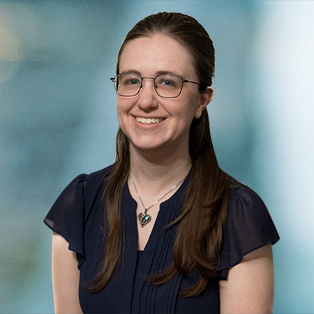 A person wearing glasses, a navy blouse, and a heart-shaped pendant necklace smiles at the camera. The background is a blurred teal and blue gradient.