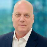 A middle-aged man with a bald head wearing a white dress shirt and dark suit jacket, posing in front of a blurred blue and green background.