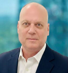 A middle-aged man with a bald head wearing a white dress shirt and dark suit jacket, posing in front of a blurred blue and green background.