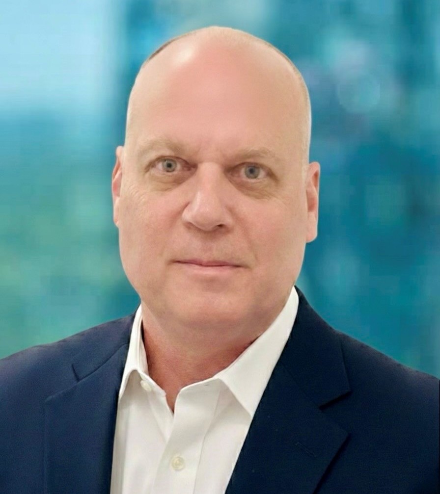 A middle-aged man with a bald head wearing a white dress shirt and dark suit jacket, posing in front of a blurred blue and green background.