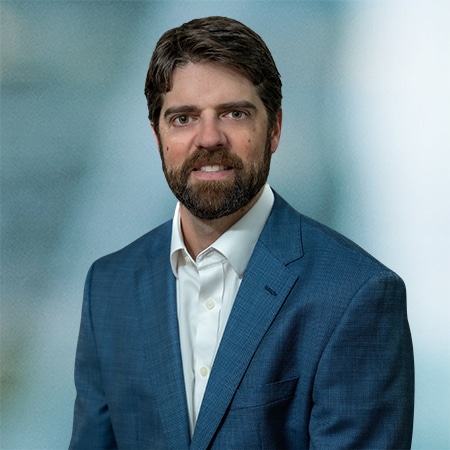 A man with dark hair and a beard, wearing a blue suit and white shirt, poses against a blurred background.
