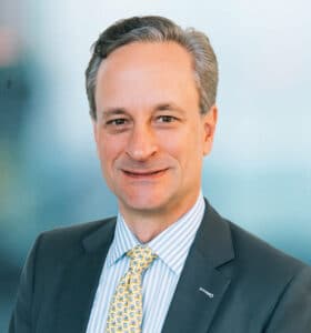A man in a suit and striped shirt, wearing a yellow tie with a subtle pattern, smiles directly at the camera. The background is softly blurred with blue tones.