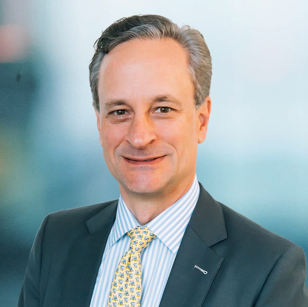 A man in a suit and striped shirt, wearing a yellow tie with a subtle pattern, smiles directly at the camera. The background is softly blurred with blue tones.