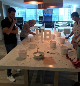 A group of people stand around a large table with buckets and scattered coins. A large HB sign is placed in the center of the table. Ceiling lights illuminate the room, which has large windows with a city view.