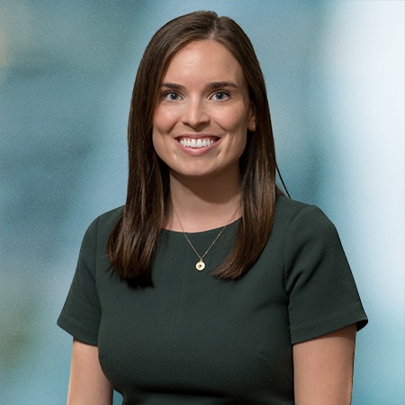 A woman with long brown hair is smiling at the camera. She is wearing a dark green dress and a round pendant necklace. The background is a soft, blurred blue and gray.