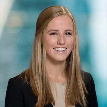 A person with long blonde hair wearing a black blazer over a beige top is smiling against a blurred blue and white background.