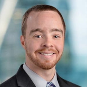A person wearing a suit and tie smiling in a professional headshot with a blurred blue and green background.