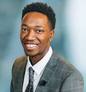 Smiling person wearing a checkered suit, white shirt, and patterned tie against a blurred background.