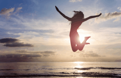 A person is joyfully jumping in the air on a beach at sunset. The silhouette is set against a vibrant sky, with the sun low on the horizon and clouds scattered above the ocean.